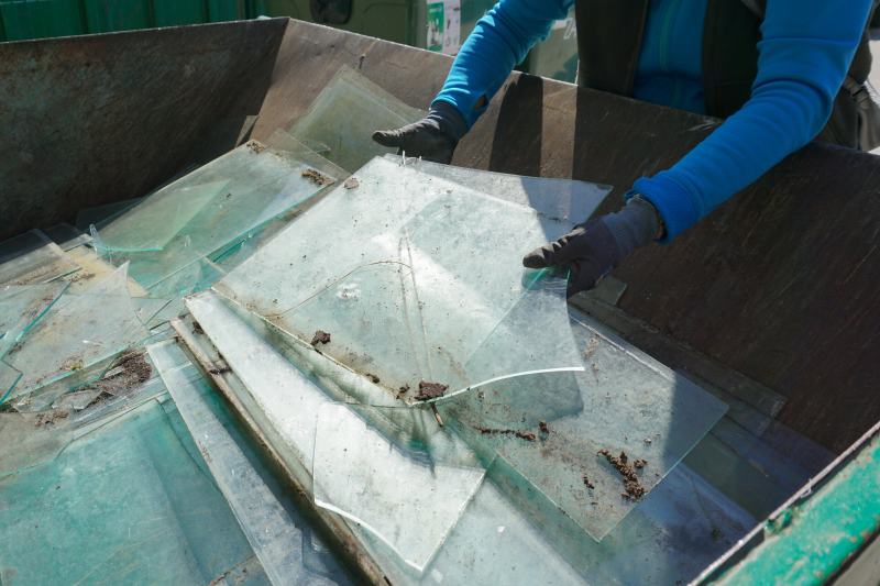 Hands in gloves disposing of old broken glass panes to waste containers in industrial recycling facility center. Processing recycled materials. Waste management, sustainability and safety