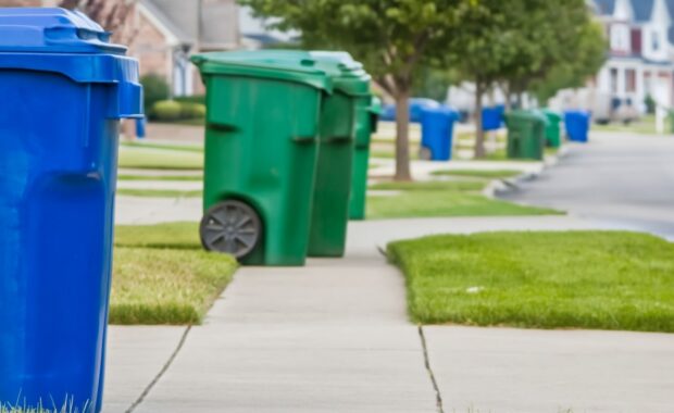 Blue and green wheeled recycling and waste bins lined up along a suburban neighborhood sidewalk on collection day