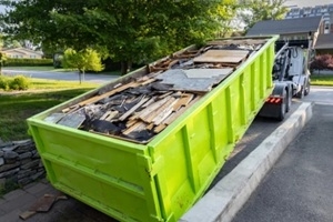 Roll-off dumpster being unloaded from a truck onto a residential driveway filled with roofing debris