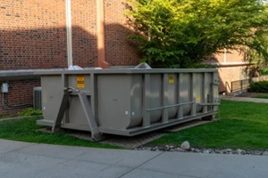 large industrial roll-off dumpster on green grass next to a brick building, ready for construction waste or demolition debris removal